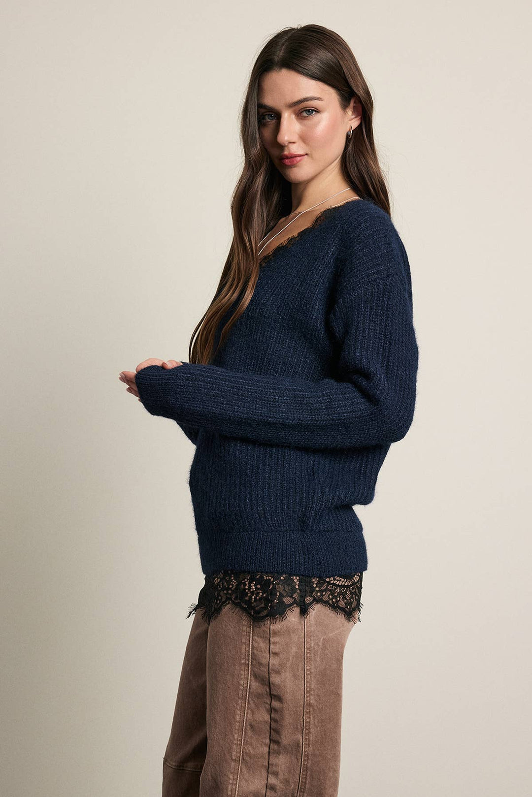 A woman modeling a dark navy blue long ribbed knit sweater with a v-neck collar, paired with brown pants and a black lace hem detail, against a plain beige studio background.