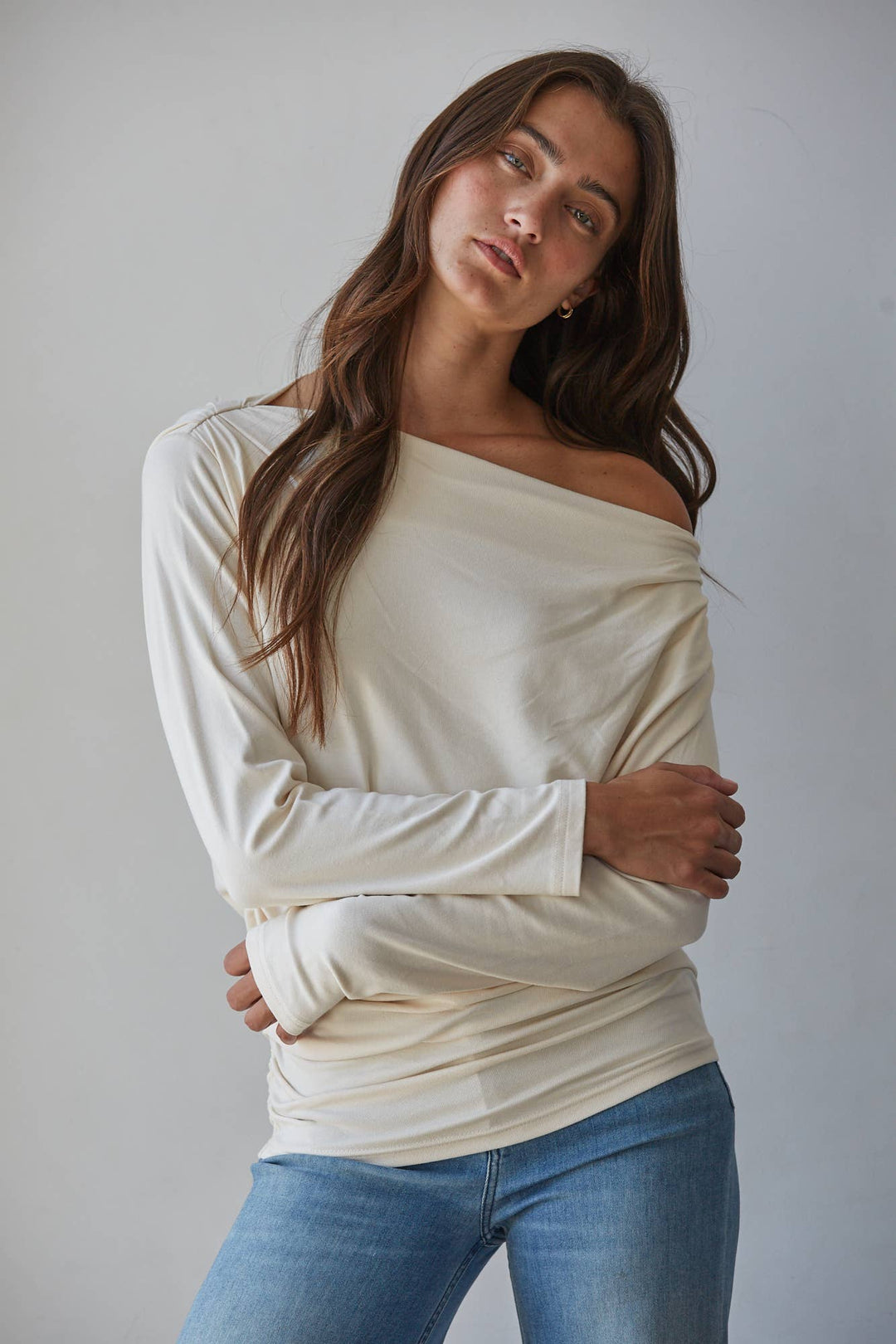A woman modeling a cream off-the-shoulder long-sleeve top with blue jeans, posed against a neutral light gray background.