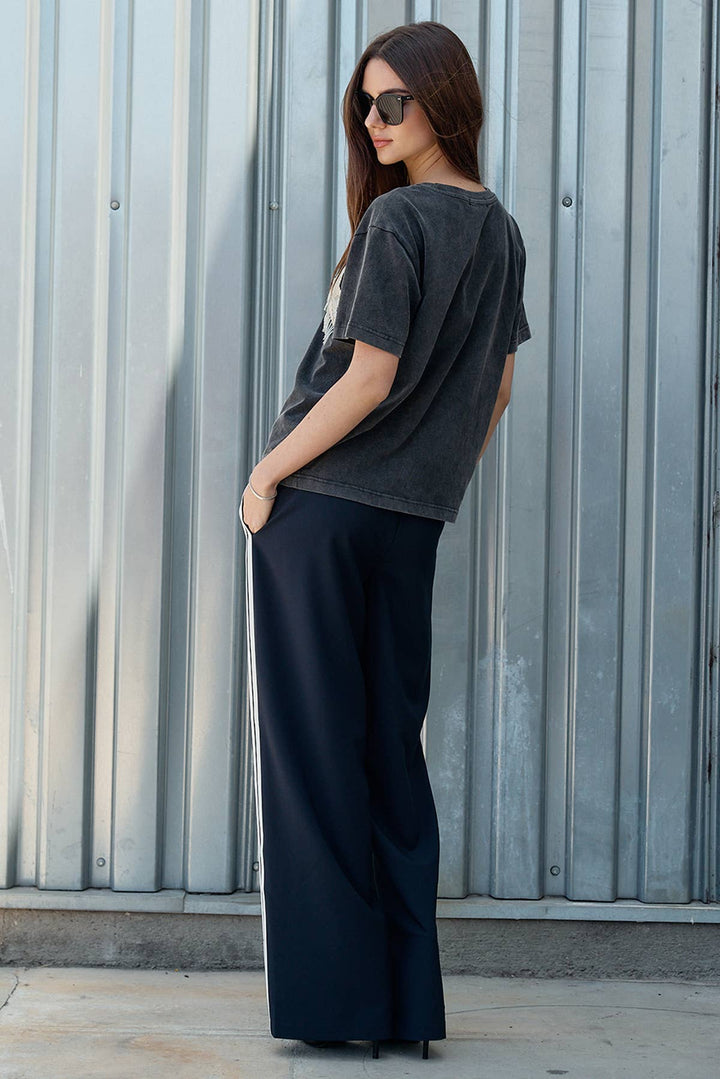 Woman wearing a dark gray t-shirt and black pants standing against a corrugated metal wall.
