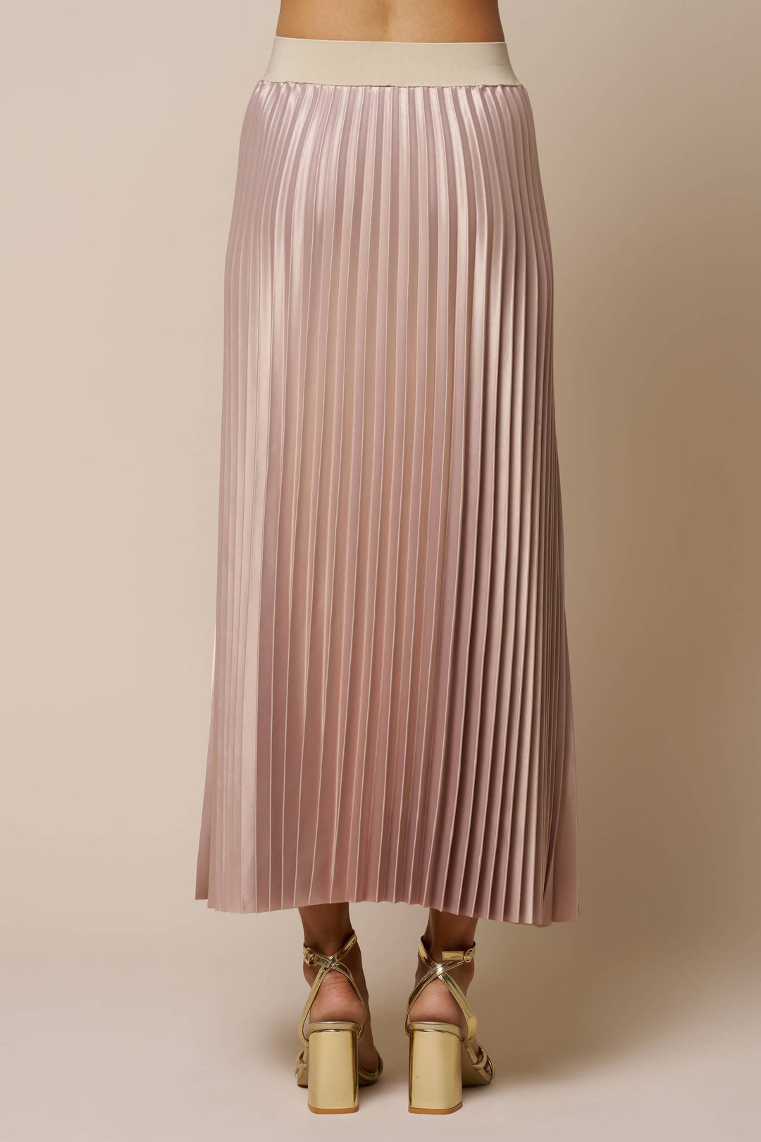 A champagne pleated midi skirt with a cream waistband, shown on a model from the back wearing gold block-heel sandals against a solid beige background.