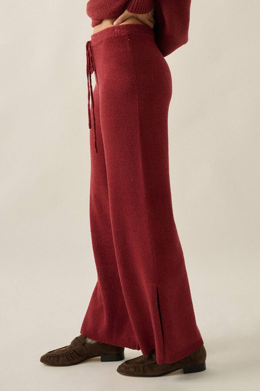 A person wearing a long, wide-leg knit sweater pants in deep burgundy red, paired with dark brown moccasin-style shoes, shown from the waist down against a plain light beige studio background.