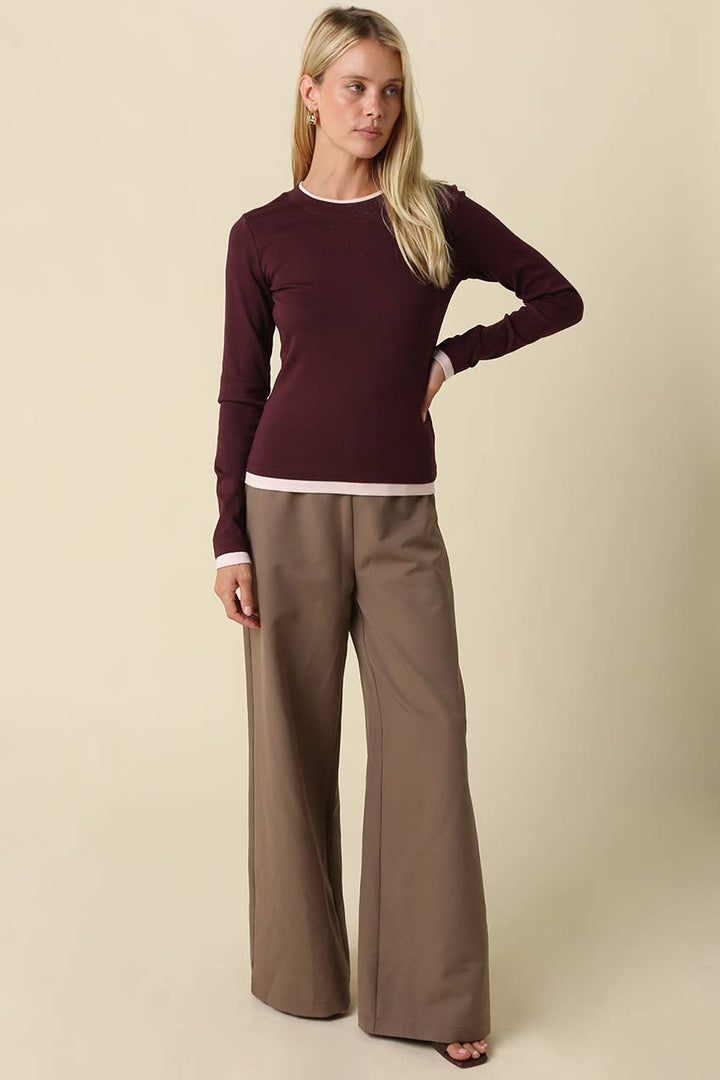 A woman modeling a long-sleeve crewneck knit top in deep burgundy with light trim, paired with wide-leg taupe pants, against a solid beige background.