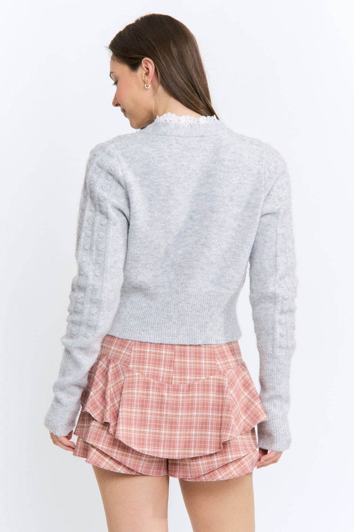 A woman shown from the back wearing a light gray ribbed knit sweater and a pink plaid ruffled mini skirt against a plain white background.