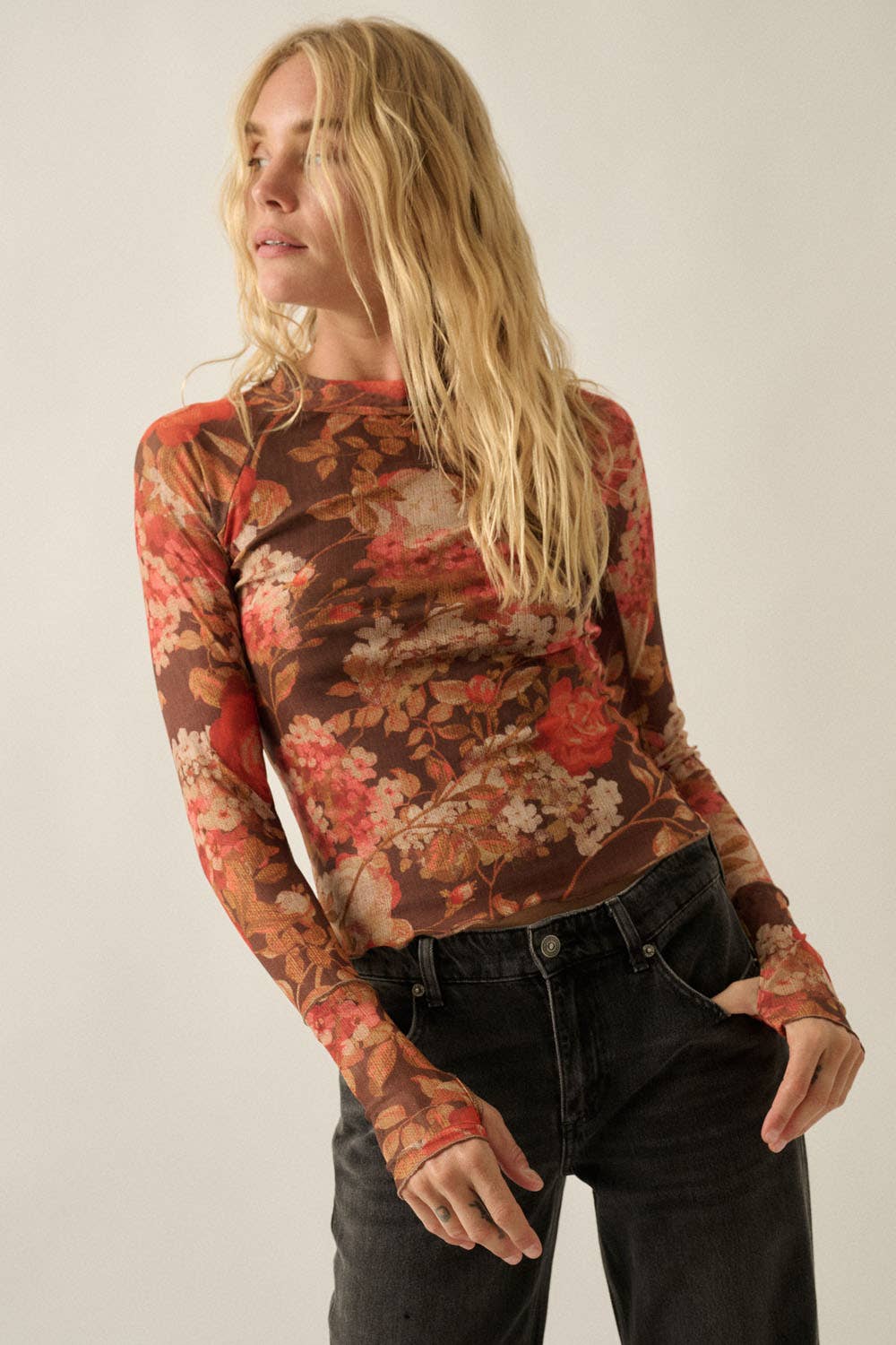 A woman wearing a long-sleeve crewneck top with a brown, red, and beige floral pattern, paired with black jeans, photographed against a plain light background.