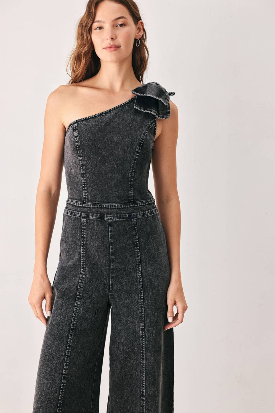 A woman modeling a dark gray washed denim one-shoulder jumpsuit with a bow shoulder detail and wide-leg pants, set against a plain light gray studio background.