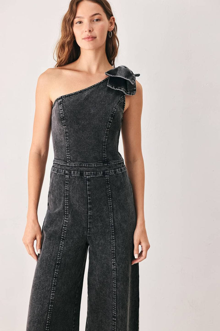 A woman modeling a dark gray washed denim one-shoulder jumpsuit with a bow shoulder detail and wide-leg pants, set against a plain light gray studio background.