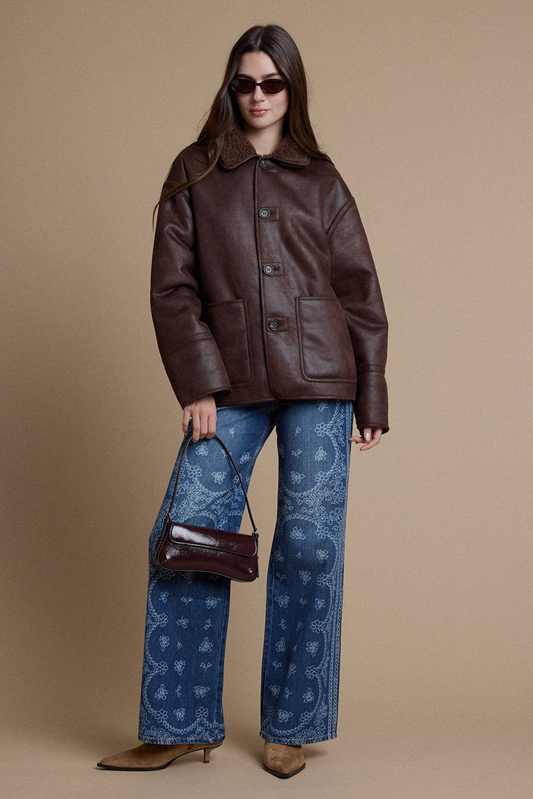 A model wearing a brown leather jacket, blue paisley-print wide-leg jeans, and brown pointed-toe heels, holding a matching brown leather handbag against a solid beige studio background.