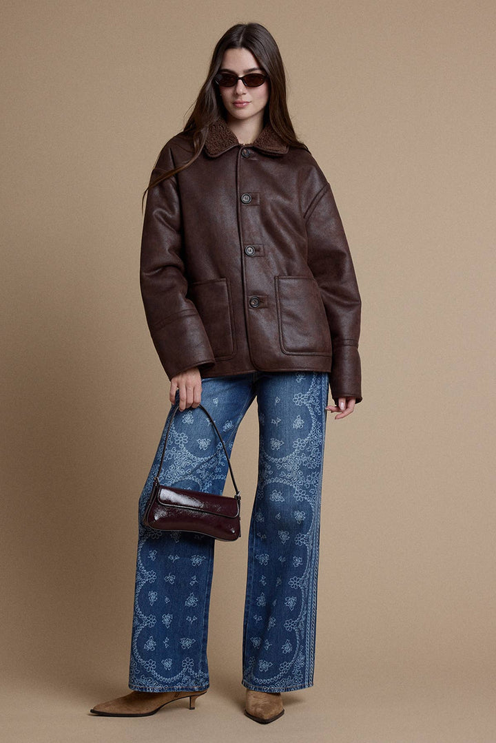 A model wearing a brown leather jacket, blue paisley-print wide-leg jeans, and brown pointed-toe heels, holding a matching brown leather handbag against a solid beige studio background.