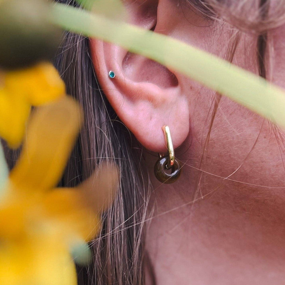 A close-up of a person's ear showcasing a gold hoop earring with a dark green teardrop-shaped stone, alongside a small green stud earring, framed by soft yellow flowers and a green leaf in the foreground.