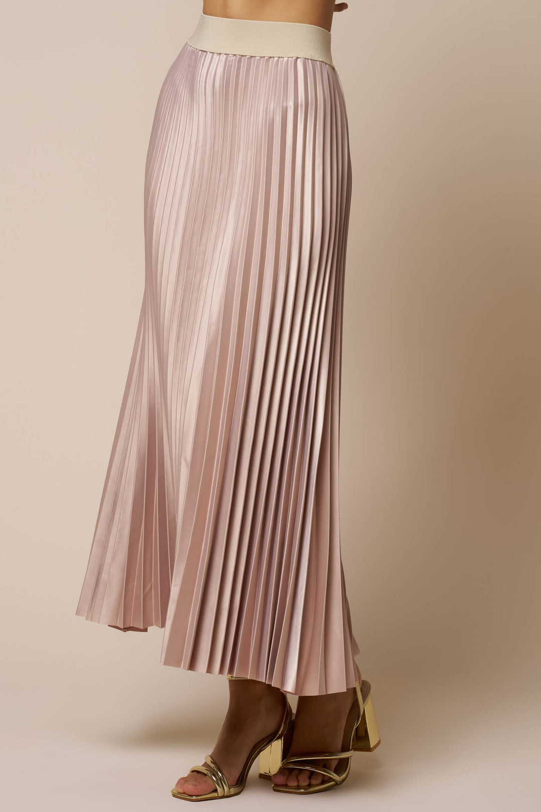 A model wearing a pleated midi skirt in a champagne color, paired with gold heeled sandals, against a solid beige studio background.
