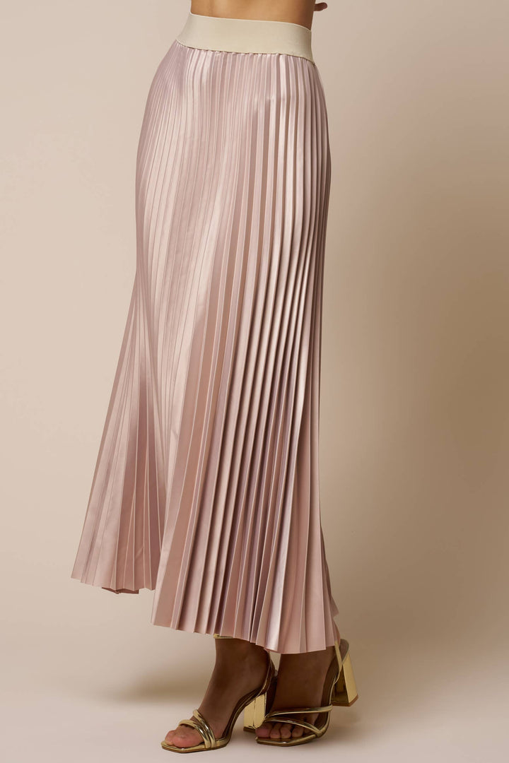A model wearing a pleated midi skirt in a champagne color, paired with gold heeled sandals, against a solid beige studio background.