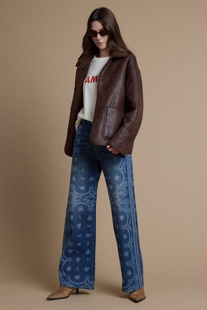 A woman models a brown leather jacket, blue paisley wide-leg jeans, a white graphic t-shirt, and brown heeled mules against a solid beige studio background.
