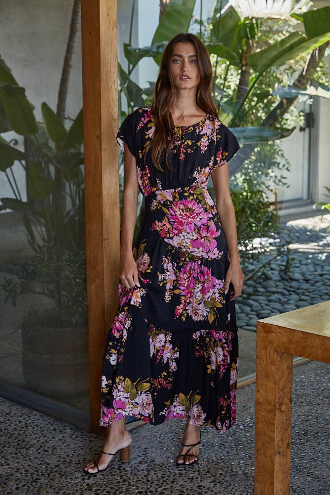 Woman wearing a black floral midi dress standing in an outdoor setting with plants and a table.