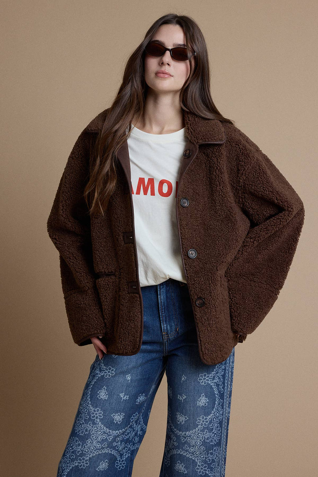 A woman modeling a brown sherpa fleece button-up jacket over a white graphic t-shirt and blue jeans with paisley patterns, set against a solid beige studio background.