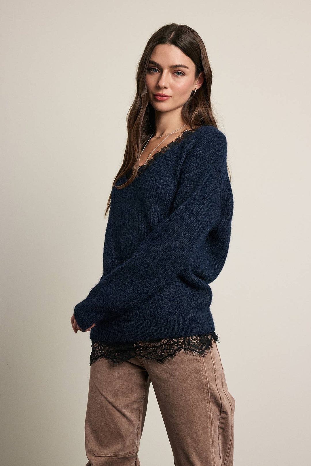 A woman modeling a dark navy blue knit sweater with a V-neck and long sleeves, styled with brown pants and a black lace hem detail, against a plain light beige studio background.