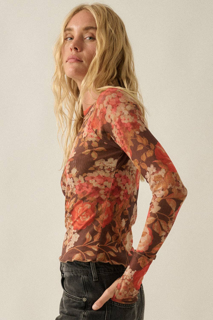 A woman wearing a long-sleeve floral print top with a brown base and red, orange, and beige floral pattern, paired with dark jeans, photographed against a plain light beige studio background.