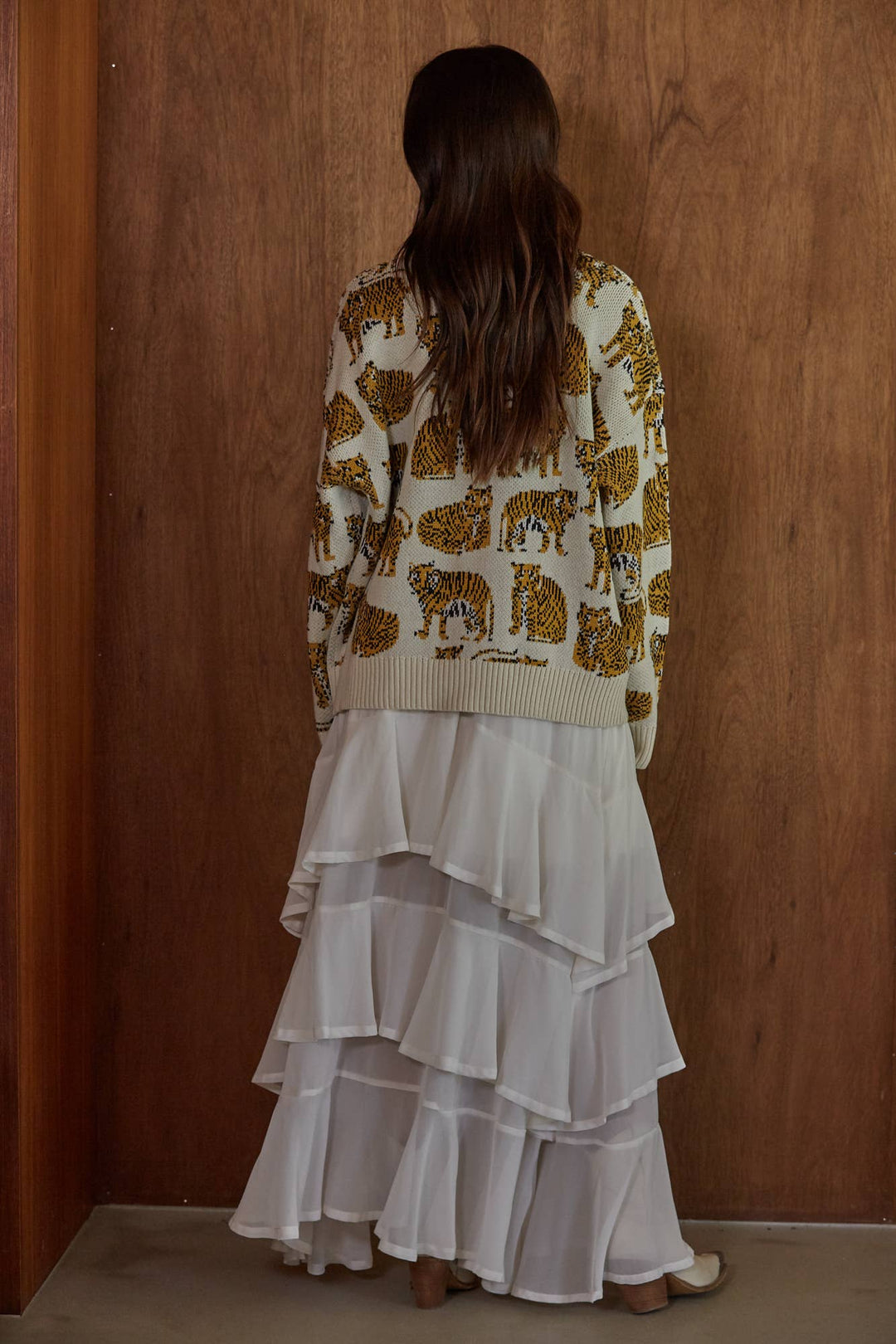 A woman viewed from behind wearing a cream knit cardigan with a brown animal print and a long white tiered skirt, standing against a wooden wall.