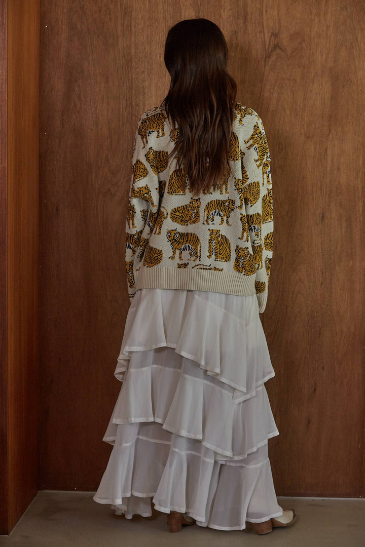 A woman viewed from behind wearing a cream knit cardigan with a brown animal print and a long white tiered skirt, standing against a wooden wall.