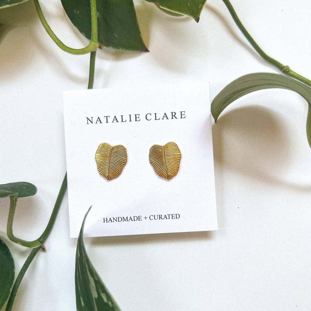 Gold leaf-shaped earrings on a 'Natalie Clare' branded card with green leaves in the background.