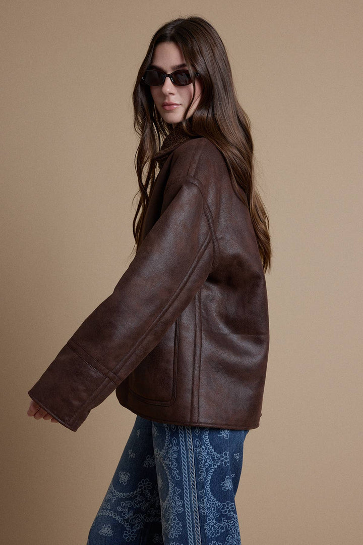 A woman wearing a dark brown faux-leather jacket and blue paisley pants, photographed against a solid beige studio background.