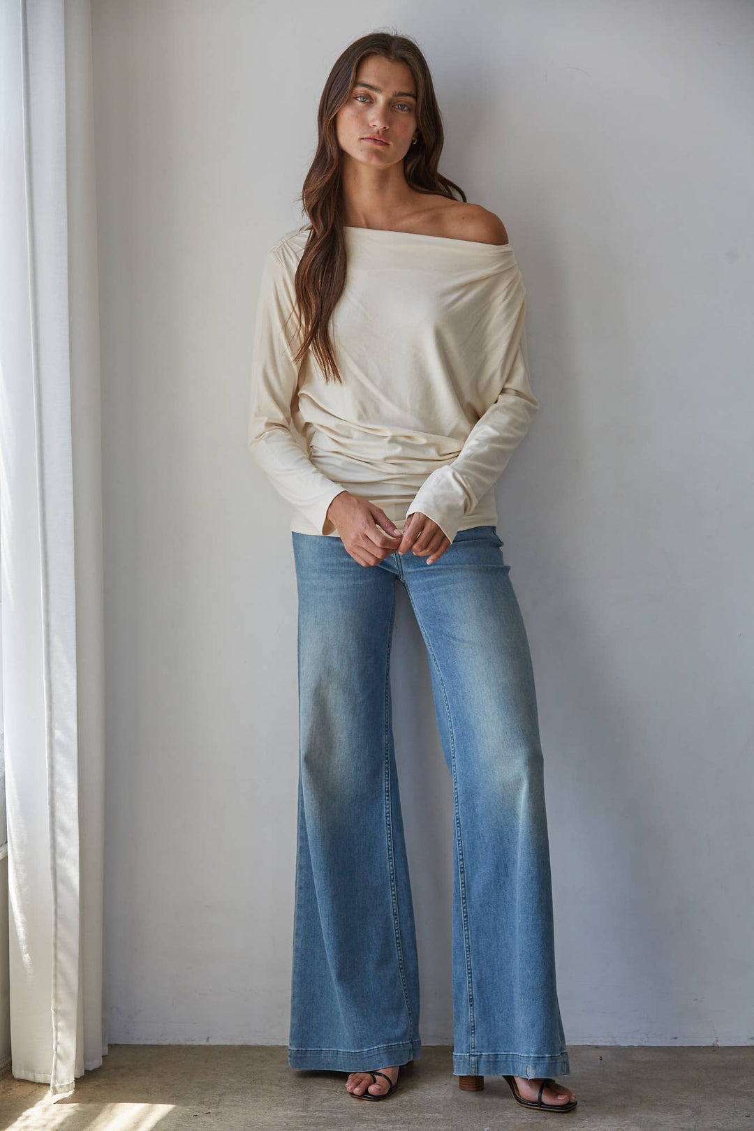 A woman stands indoors against a light gray wall wearing a cream off-the-shoulder knit long-sleeve top and light blue wide-leg jeans, with brown sandals.