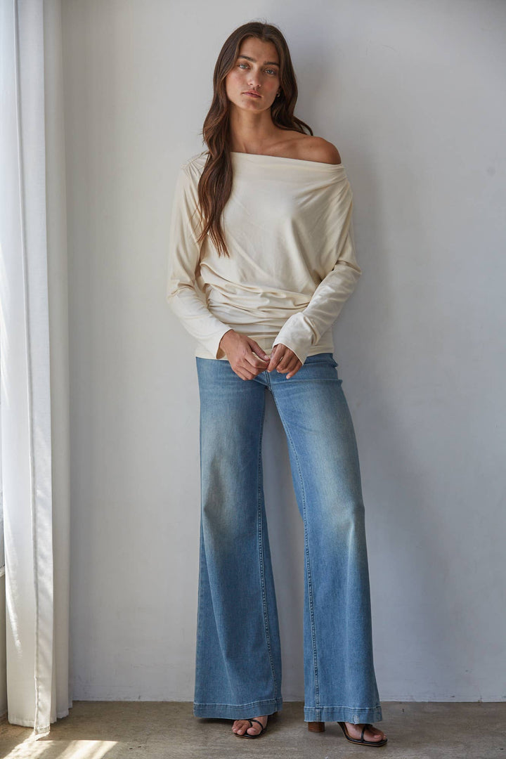 A woman stands indoors against a light gray wall wearing a cream off-the-shoulder knit long-sleeve top and light blue wide-leg jeans, with brown sandals.