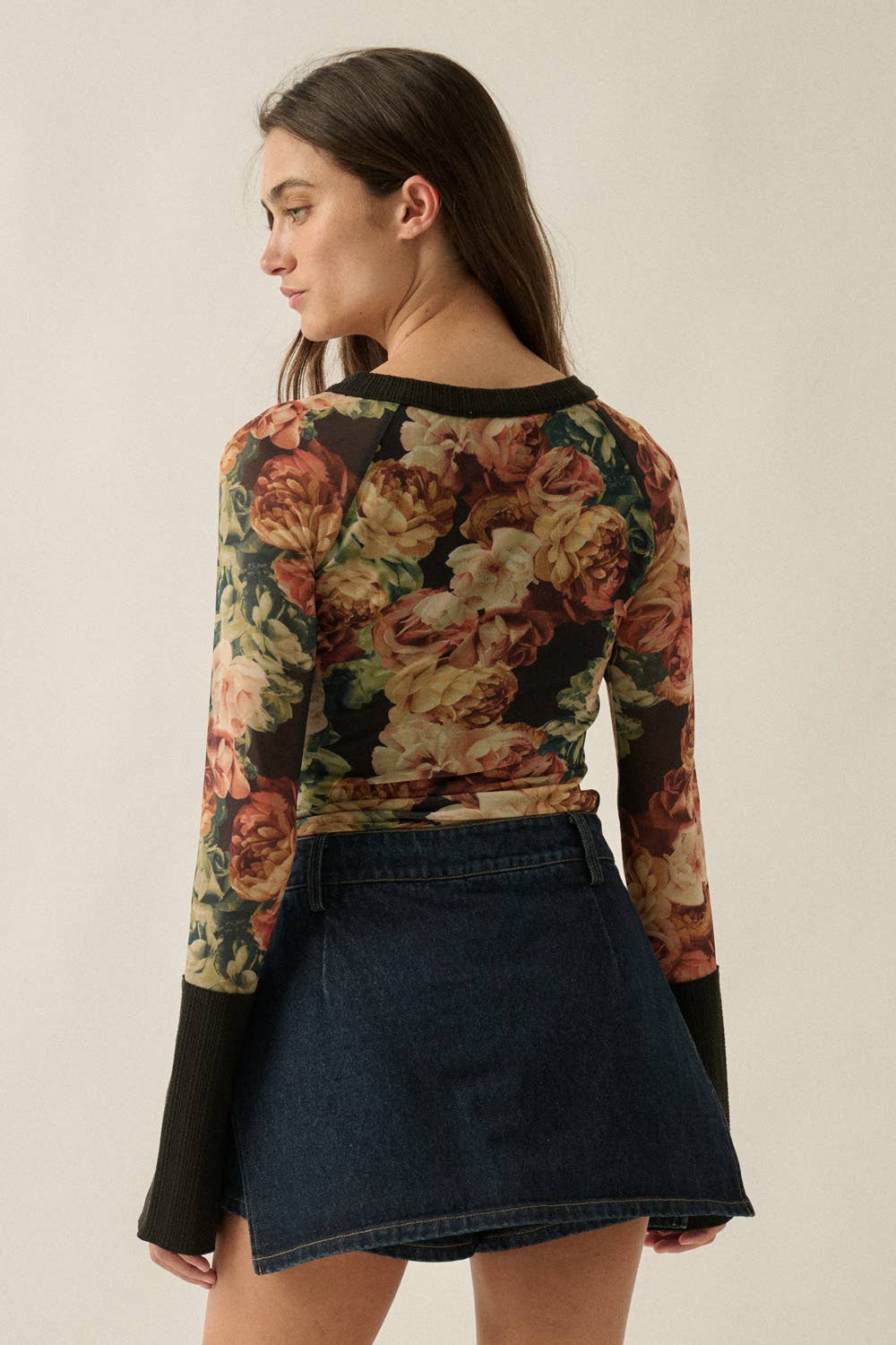 A woman shown from the back wearing a long-sleeve floral top with black cuffs and a dark blue denim mini skirt, against a plain light beige studio background.