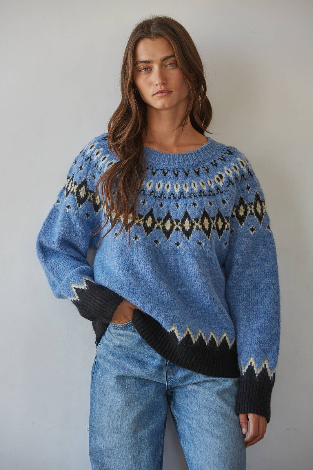 A woman wearing a blue knit sweater with a black and cream Fair Isle pattern across the chest and cuffs, paired with light-wash blue jeans, standing against a plain light gray wall.