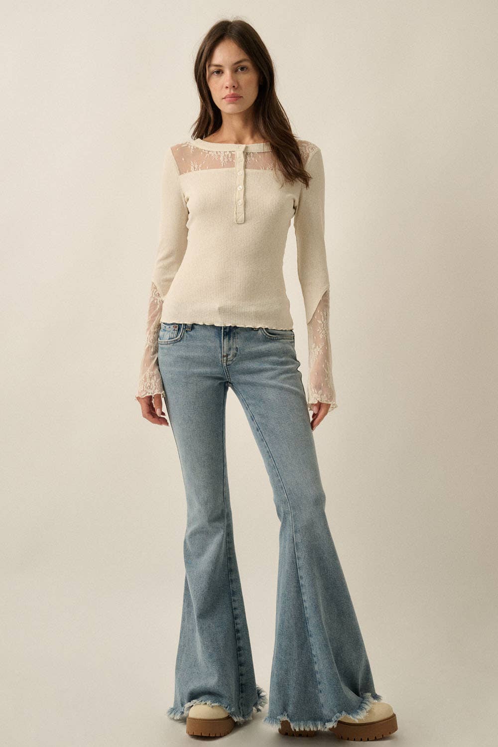 A woman modeling a cream ribbed knit long-sleeve top with sheer lace panels at the neckline and cuffs, paired with light blue flared jeans and beige platform shoes, against a plain beige studio background.