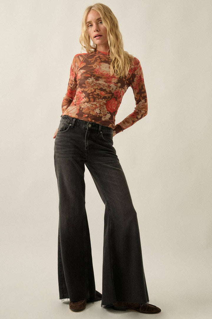 A woman modeling a long-sleeve mock-neck top with a rust-orange and brown abstract floral pattern, paired with dark gray wide-leg jeans and brown slip-on shoes, against a plain light beige studio background.
