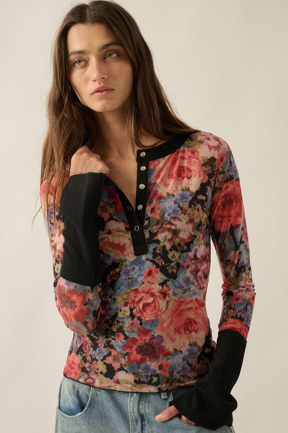 A woman wearing a long-sleeve floral henley top with black ribbed cuffs and collar, paired with light blue jeans, against a plain light beige background.
