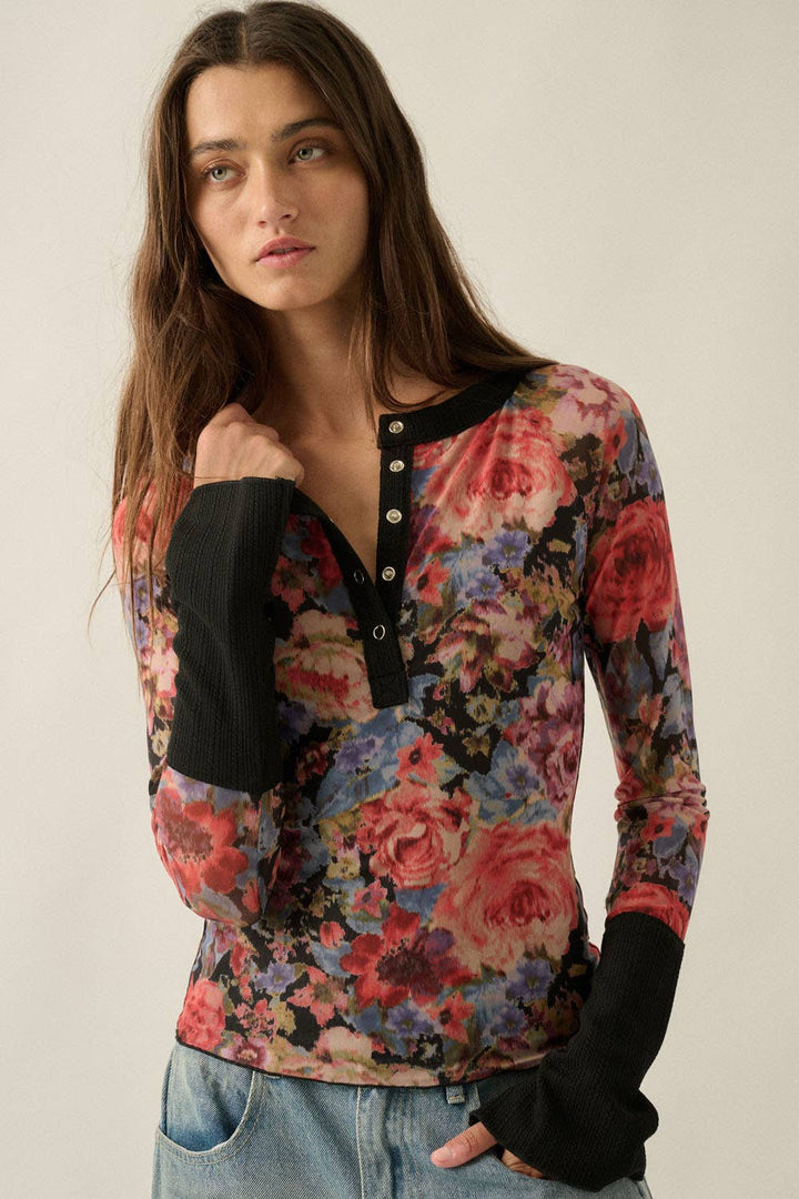 A woman wearing a long-sleeve floral henley top with black ribbed cuffs and collar, paired with light blue jeans, against a plain light beige background.