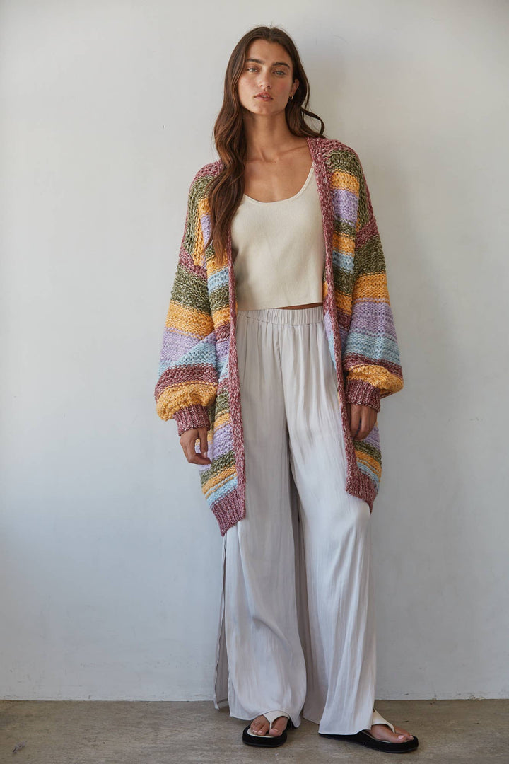 A woman stands against a plain light gray wall wearing a long, open-front chunky knit cardigan with horizontal multicolor stripes in pink, purple, blue, yellow, green, and orange, over a cream tank top and white wide-leg pants.