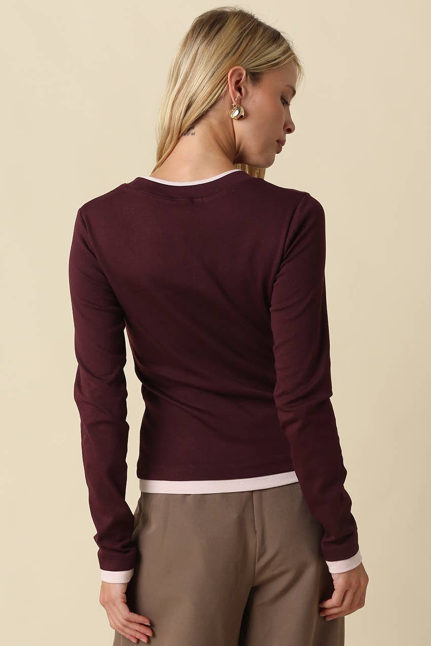 A woman shown from the back wearing a long-sleeve crewneck knit top in deep burgundy with white ribbed trim at the collar, cuffs, and hem, paired with taupe pants, against a solid beige background.