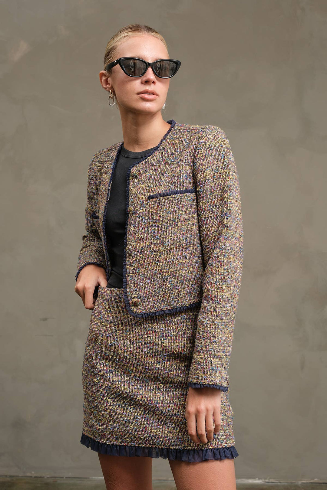 A woman modeling a tweed mini skirt suit with a matching cropped jacket, featuring a multicolor blend of beige, brown, and blue with dark navy piping, styled with a black top and black sunglasses against a textured gray wall.