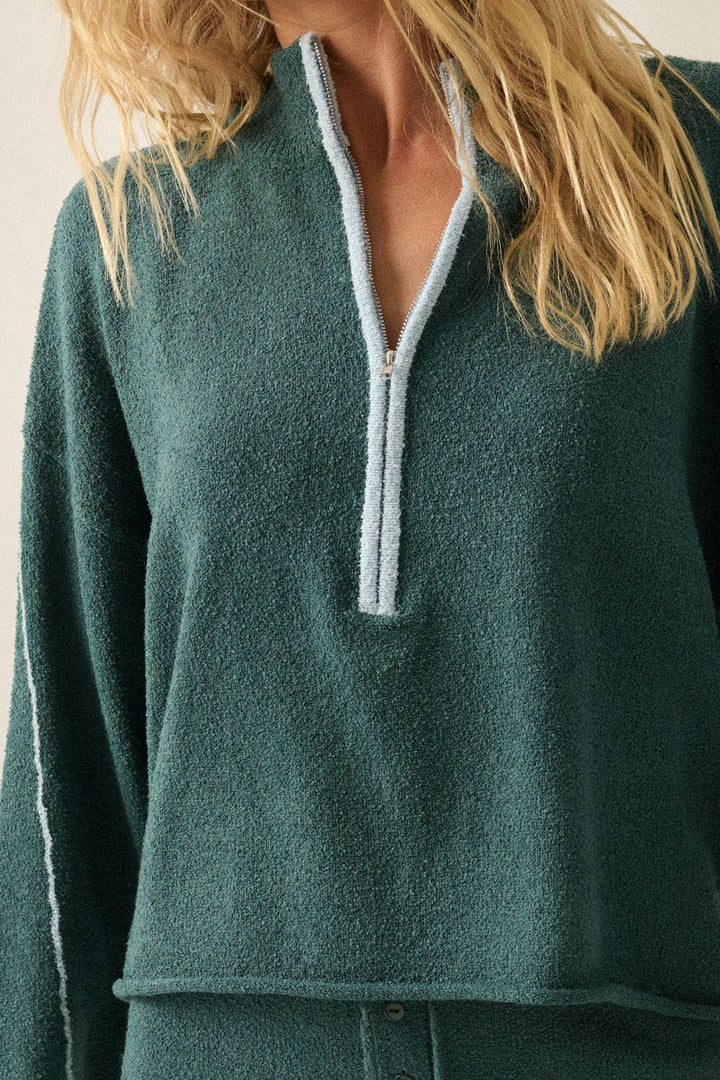 A close-up of a woman wearing a dark teal green quarter-zip pullover sweater with light blue-gray contrast trim along the zipper and sleeve.