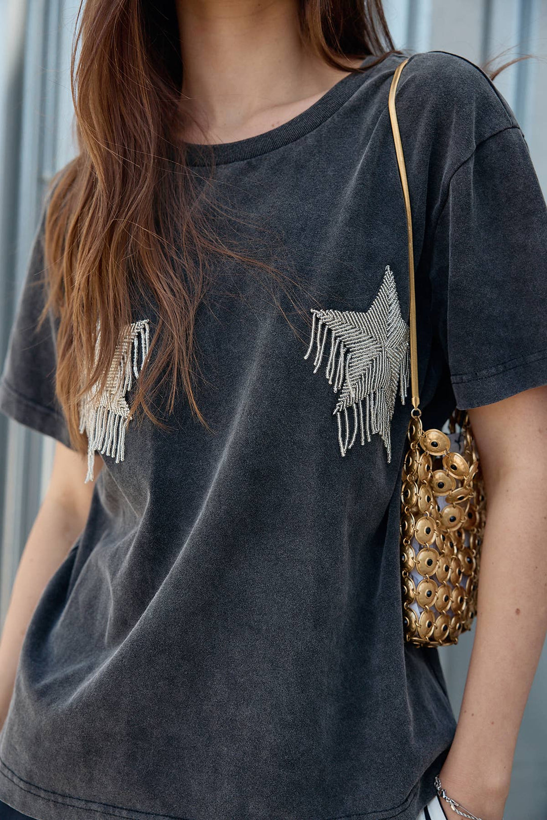 Person wearing a dark gray t-shirt with fringe details and carrying a gold handbag.