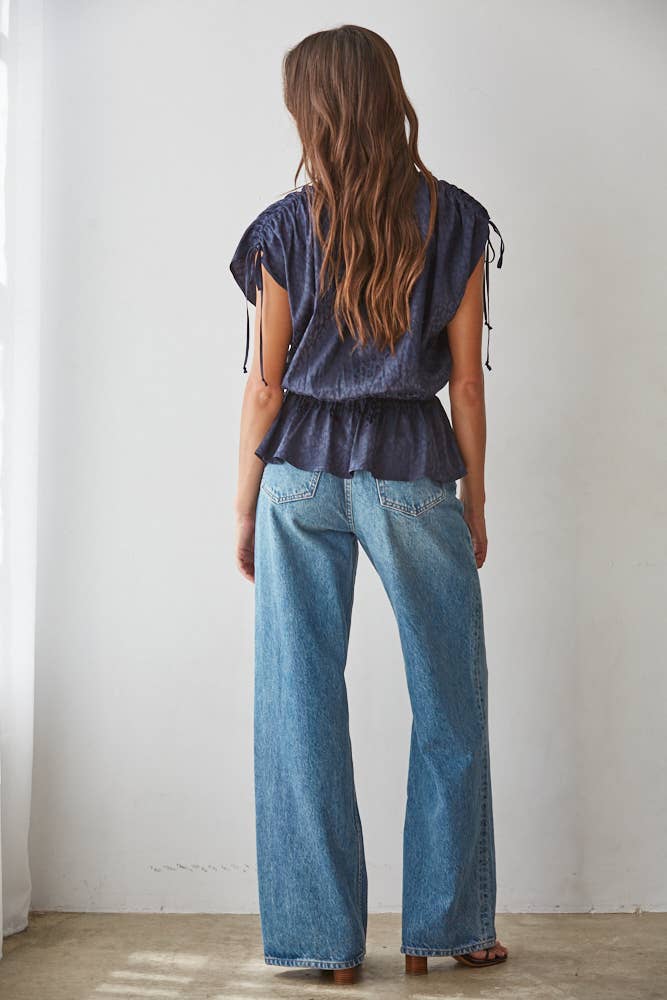 A woman viewed from behind wearing a dark blue ruffled sleeveless top and light blue wide-leg jeans, standing against a plain white wall.