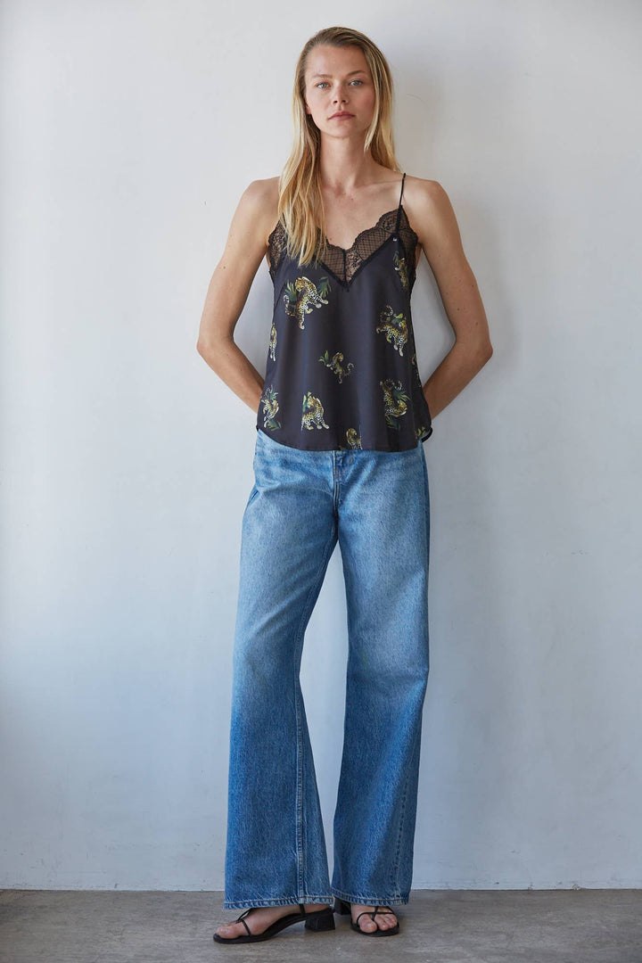 A woman stands against a light gray wall wearing a dark camisole top with a animal print and lace trim, paired with wide-leg blue jeans and black sandals.