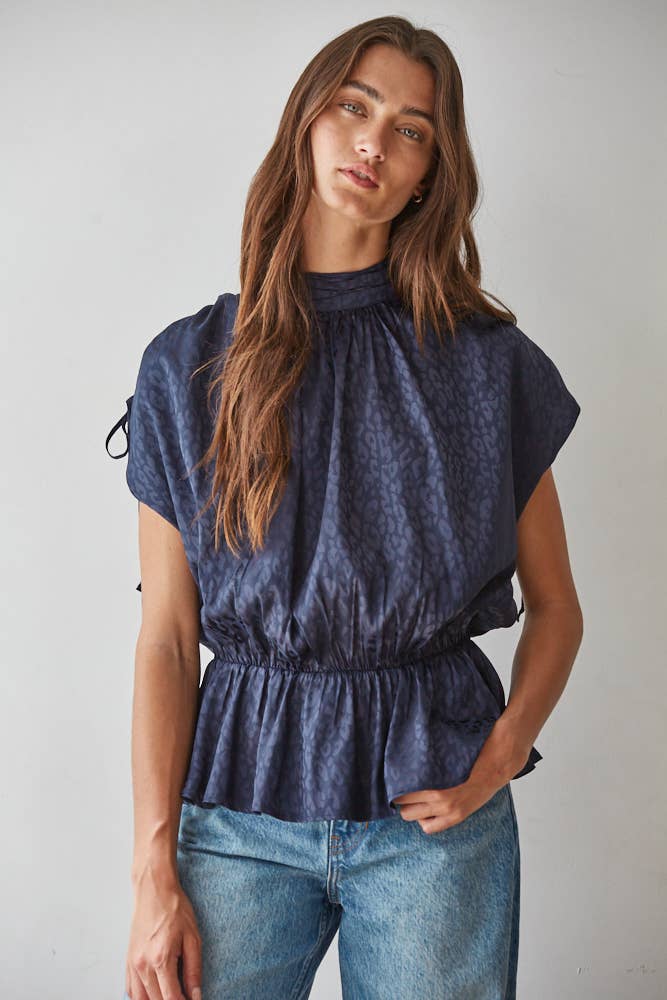 A woman wearing a dark blue patterned short-sleeve blouse with a gathered peplum waist and blue jeans, standing against a plain light gray background.