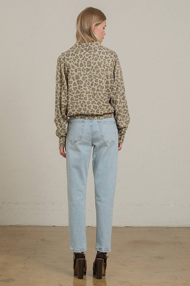 A woman is shown from behind wearing a beige and brown leopard print long-sleeve shirt, light blue jeans, and brown block-heel boots, standing against a neutral off-white wall.