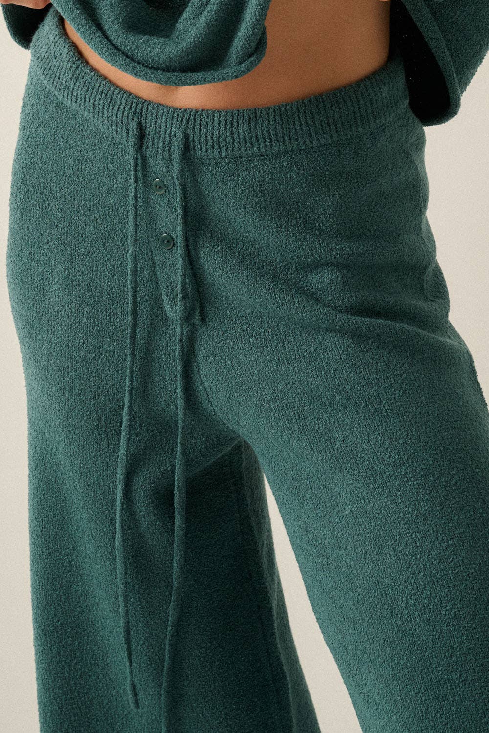 Close-up of a woman wearing dark teal knit pants with a drawstring waist and front button detail, paired with a matching top.