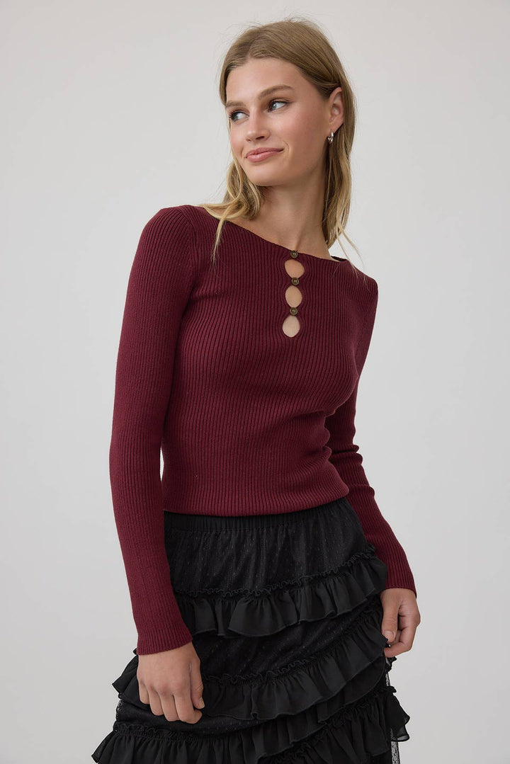 A woman wearing a ribbed long-sleeve top in deep wine color with three large decorative buttons at the neckline, paired with a black ruffled skirt, against a plain light gray studio background.