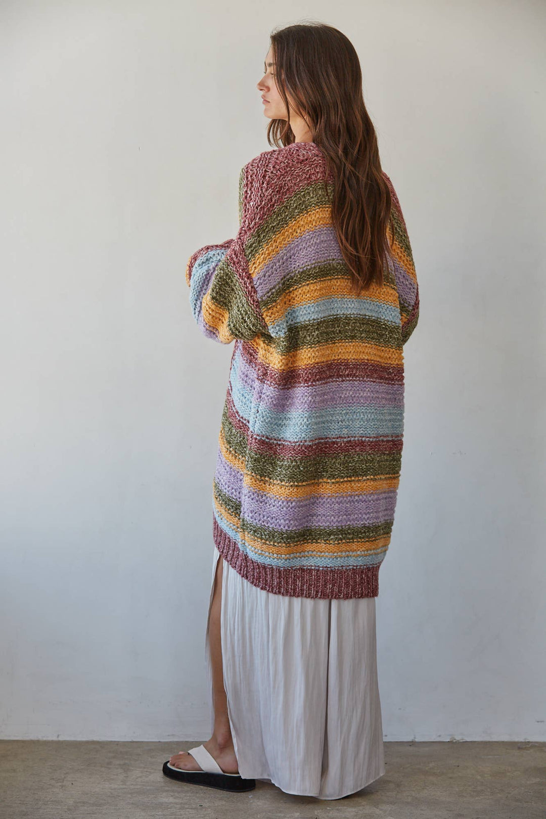 A woman stands with her back to the camera wearing a loose, oversized striped knit cardigan in pastel rainbow colors over a long white skirt.