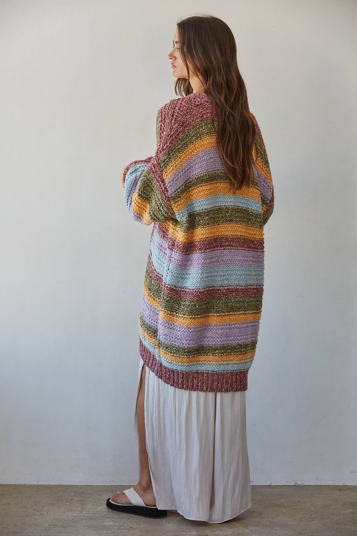 A woman stands with her back to the camera wearing a loose, oversized striped knit cardigan in pastel rainbow colors over a long white skirt.