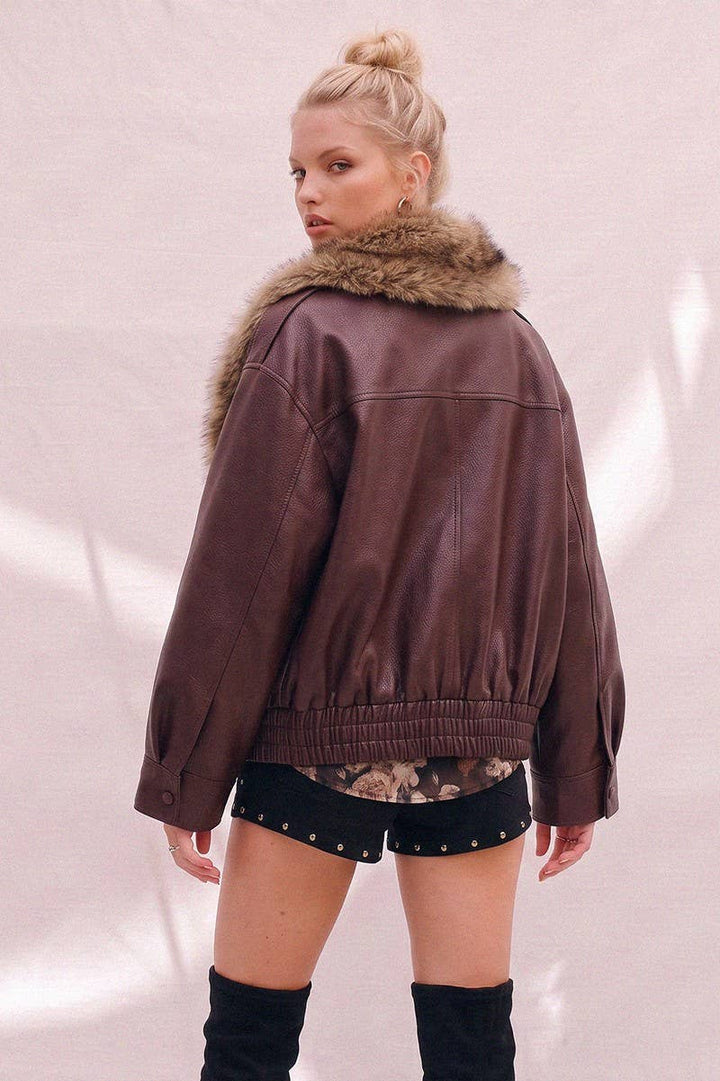 A woman wearing a burgundy faux-leather bomber jacket with a detachable brown faux-fur collar, paired with black studded shorts and black over-the-knee boots, photographed against a light pink textured backdrop.