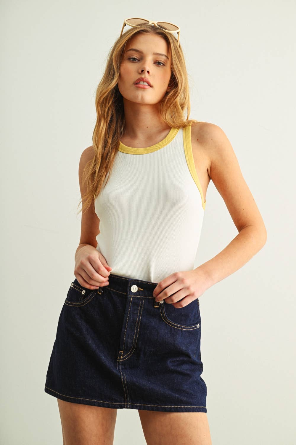 Woman wearing a white tank top with yellow trim and a dark denim skirt on a plain background