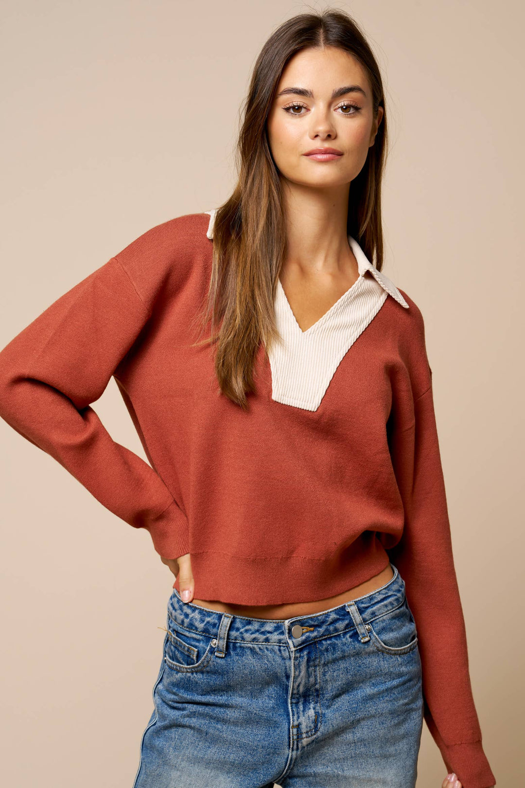 A woman wearing a rust-colored knit sweater with a cream V-neck collar, paired with blue jeans, against a solid beige background.