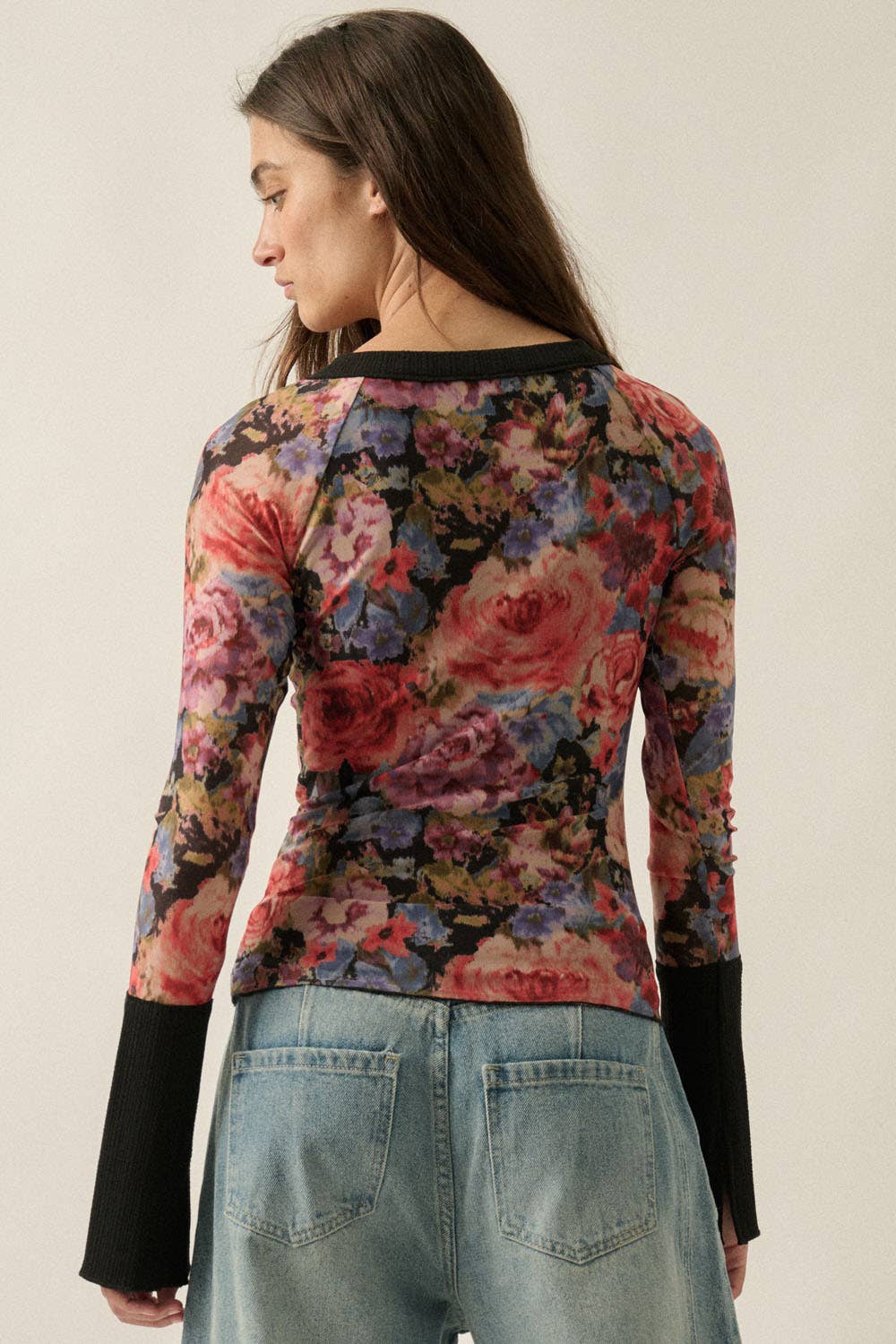 A woman shown from the back wearing a fitted long-sleeve mesh top with a dense floral print in pink, red, blue, and purple tones over a black base, paired with light blue jeans against a plain light beige studio background.