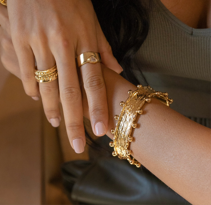 Close-up of a hand wearing gold rings and a bracelet on a neutral background